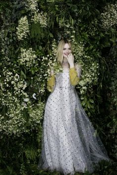 a woman in a white dress is surrounded by flowers and greenery with her hands to her face