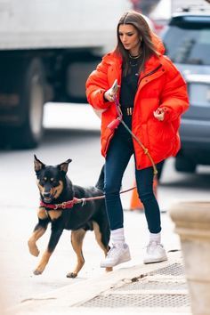 a woman in an orange jacket walking her dog on a leash and looking at the camera