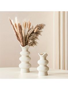 two white vases with dried plants in them sitting on a table next to each other