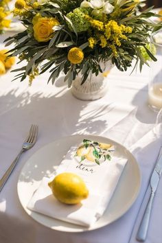 a white plate topped with lemons next to a vase filled with yellow flowers and greenery