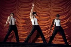 three men in white shirts and black pants on stage with their hands up to the side