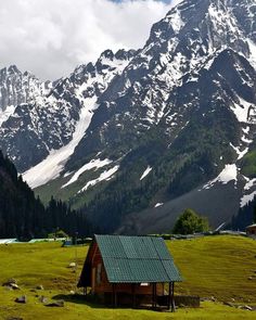 a small cabin in the middle of a field with snow covered mountains behind it,