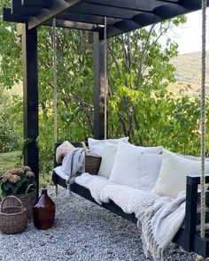 a porch swing with pillows and blankets on it, in front of some trees outside