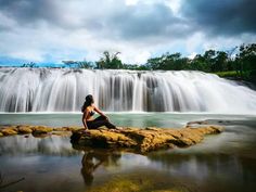 a woman sitting on top of a rock next to a waterfall in the middle of a body of water