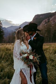 a bride and groom kissing in front of mountains with flowers on their wedding day, wearing cowboy hats