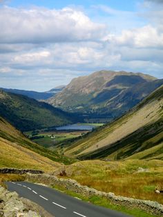 an empty road in the middle of mountains with grass on both sides and water at the bottom