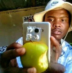 a man is holding an apple and taking a selfie with his cell phone while wearing a hat