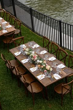 an outdoor table set up with place settings and flowers on it, next to the water