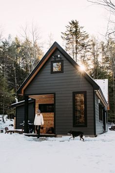 a woman standing in front of a small house with a dog on the snow covered ground