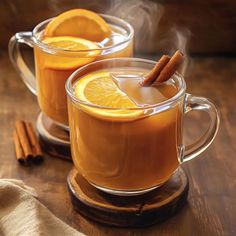 two mugs filled with hot drinks and cinnamon on top of a wooden table next to an orange slice