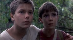 two young boys standing next to each other in front of some trees and bushes with one looking at the camera