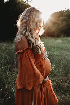 a pregnant woman in an orange dress standing in tall grass with the sun shining behind her