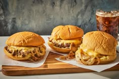 three cheeseburgers sitting on top of a wooden cutting board next to a drink