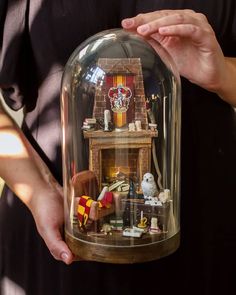 a person holding a fake fireplace under a clochet glass dome with miniature figurines in it