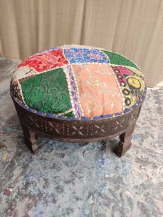 a patchwork ottoman sitting on top of a stone floor next to a wall with a curtain in the background