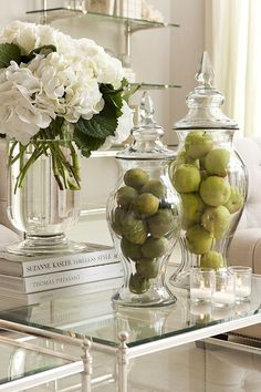 a glass table topped with two vases filled with green apples and white hydrangeas