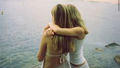 two young women standing next to each other near the water with their arms around each other