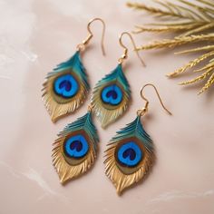 three pairs of blue and gold peacock feather earrings on top of a pink surface next to a plant