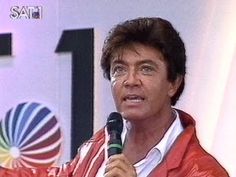 a man holding a microphone in his right hand and wearing a red jacket on the left