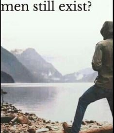 a man standing on top of a log next to a body of water with mountains in the background