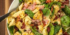 a white bowl filled with pasta and spinach on top of a wooden table next to a fork