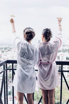 two women in robes standing on a balcony with their arms raised and holding wine glasses