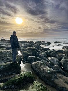 a man standing on top of a rocky beach next to the ocean under a cloudy sky