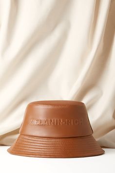 A caramel-brown bucket hat embossed with “Melanin-Rich”. Cheap Trendy Brown Bucket Hat, Luxury Bucket Hat With Curved Brim For Vacation, Luxury Brimmed Bucket Hat For Summer, Luxury Woven Bucket Straw Hat, Luxury Flat Brim Bucket Hat For Summer, Luxury Cotton Hats, Luxury Short Brim Cotton Hat, Luxury Adjustable Cotton Hats, Luxury Trendy Bucket Hat With Short Brim