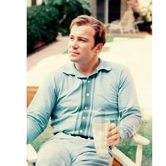 a man sitting in a chair holding a drink and looking off into the distance while wearing a blue shirt