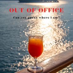 an orange drink sitting on top of a wooden table next to the ocean with text out of office can you guess where i am?