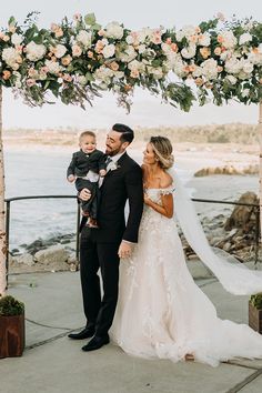 a bride and groom holding their son under an arch with flowers on the beach in front of them