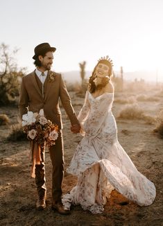 a bride and groom holding hands walking through the desert at sunset in their wedding attire