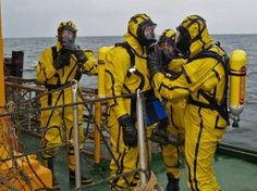 three people in yellow suits standing on a boat near the ocean and wearing gas masks