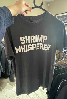 a person holding up a shirt that says shrimp whisperer on it in front of some clothes