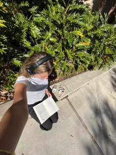 a woman sitting on the sidewalk with an open book and headphones in her ears