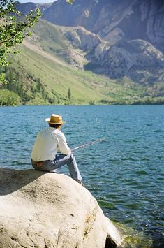 a man sitting on top of a rock next to a body of water with mountains in the background