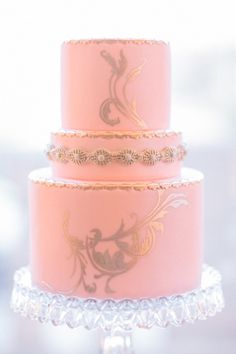 a three tiered pink cake with gold designs on the top and bottom, sitting on a glass stand