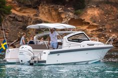 three people on a white boat in the water near some cliffs and trees, with one person standing at the front of the boat