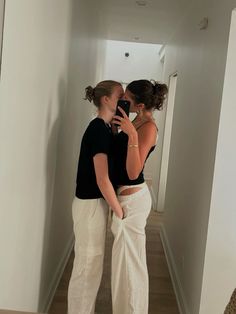 two women standing in a hallway looking at their cell phones while they both wear white pants and black tops