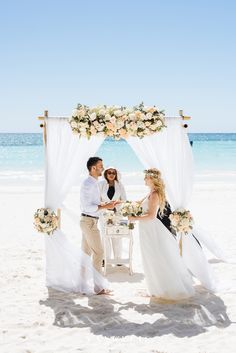 a couple getting married on the beach in front of an arch decorated with flowers and greenery