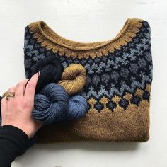 a hand holding yarn next to a knitted sweater on top of a white table