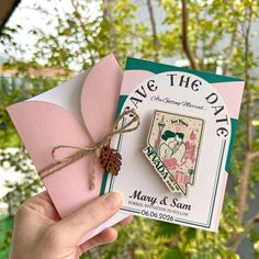 someone holding up a pink and green save the date card with a pine cone on it