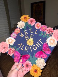 someone holding up a graduation cap with flowers and butterflies on it that says we'll be alright