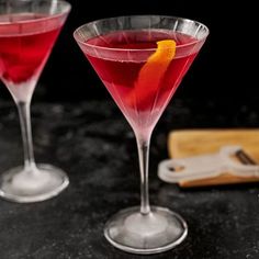 two wine glasses with red liquid and an orange peel in them on a black table