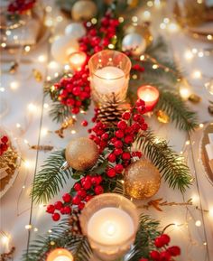 a table with candles and christmas decorations on it, including red berries, pine cones, evergreen leaves and gold baubs