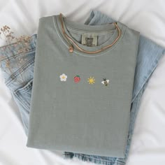 an embroidered t - shirt is laying on top of some blue jean pants and flowers