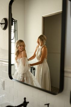 two women in wedding dresses looking at each other through a bathroom mirror with the reflection of them