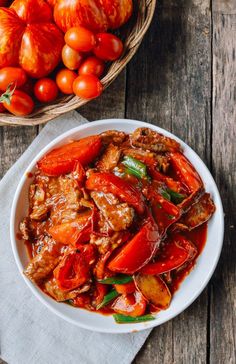 a white bowl filled with meat and vegetables next to a basket of tomatoes on a wooden table