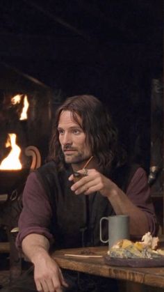 a man with long hair sitting at a table in front of a fire place holding a cup