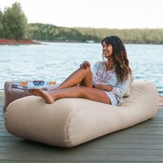 a woman sitting on top of a bean bag chair next to the water with a tray of food